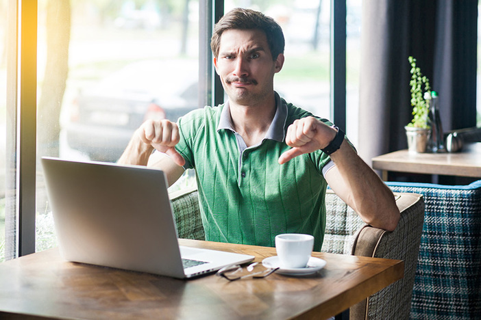 5 Ways to Lose Your Wholesale Deal (After Signing the Contract) Dislike! Young dissatisfied businessman in green t-shirt sitting, working on laptop, looking at camera and showing thumbs down. business and freelancing concept. indoor shot near big window at daytime