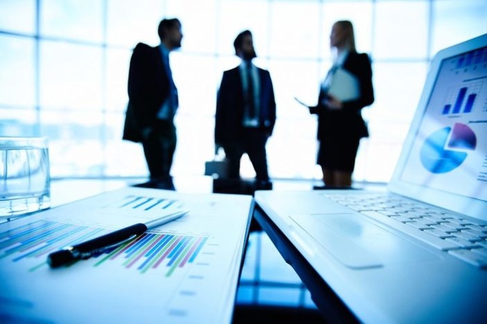 Mastering These 2 Skills Is Critical to Success in Real Estate Investing brightly blue lit room, silhouettes of two men and a woman wearing business attire talking in background, spreadsheets, graphs, pen, laptop on boardroom table in foreground