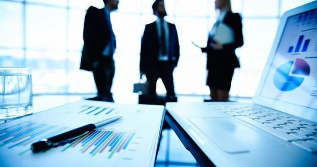 Mastering These 2 Skills Is Critical to Success in Real Estate Investing brightly blue lit room, silhouettes of two men and a woman wearing business attire talking in background, spreadsheets, graphs, pen, laptop on boardroom table in foreground