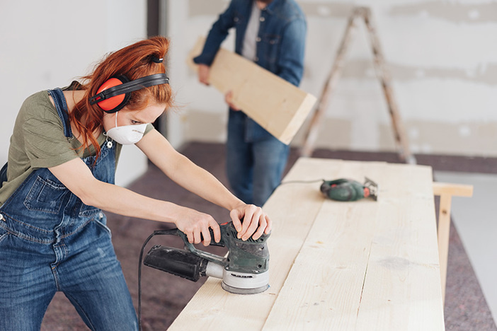 Investor Spotlight: From Accidental House Hack to Financial Freedom Featuring Whitney Hutten young woman sanding planks of wood with an orbital sander in an unfinished room during DIY renovations